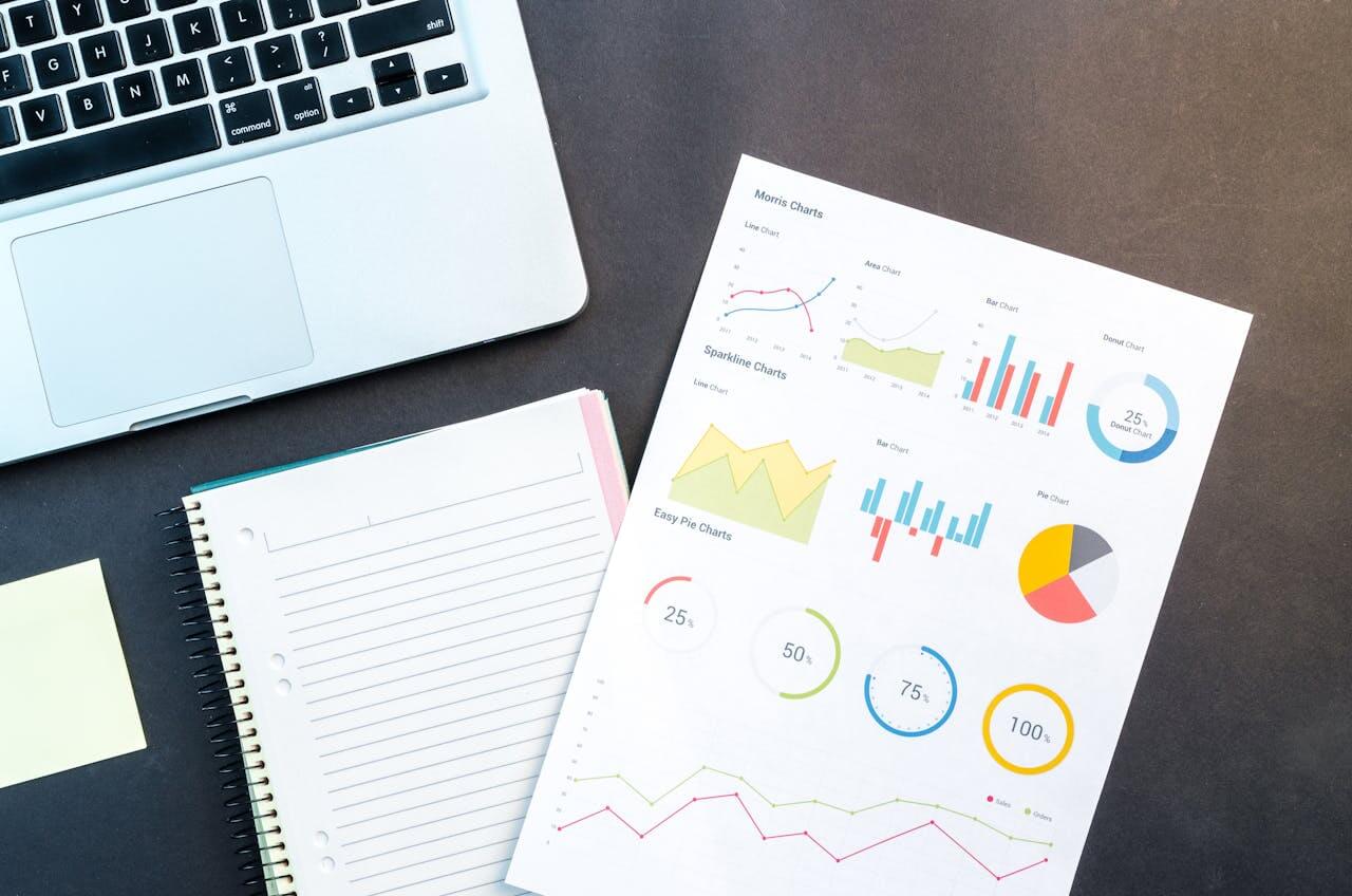 Auf dem Tisch stehen ein Laptop, ein Spiralblock und eine Seite voller bunter Grafiken und Diagramme. Diese Einrichtung spiegelt den typischen Arbeitsplatz von BrandSimpli wider, wo Kreativität und strategisches Denken mühelos zusammenkommen.