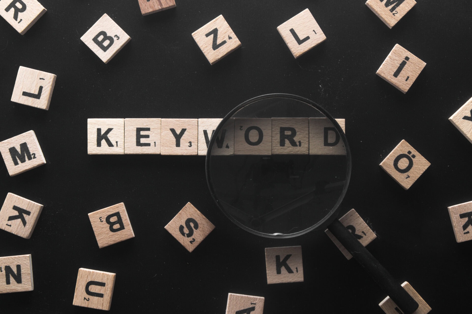 Auf einer schwarzen Fläche sind Scrabble-Steine mit dem Wort "KEYWORD" ordentlich aufgereiht. Um sie herum sind verstreute Steine und eine Lupe, die die Suche nach einem besseren Suchmaschinen-Ranking symbolisiert.