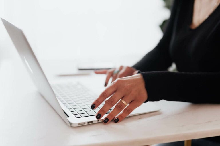 Eine Person tippt akribisch auf einem silbernen Laptop und arbeitet möglicherweise an einer ansprechenden SEO-Strategie. Die Szene spielt sich auf einem makellos weißen Tisch ab. Ihre Hände sind mit schwarzem Nagellack verziert, und ein stilvoller Ring verleiht dem digitalen Arbeitsplatz dezentes Flair.