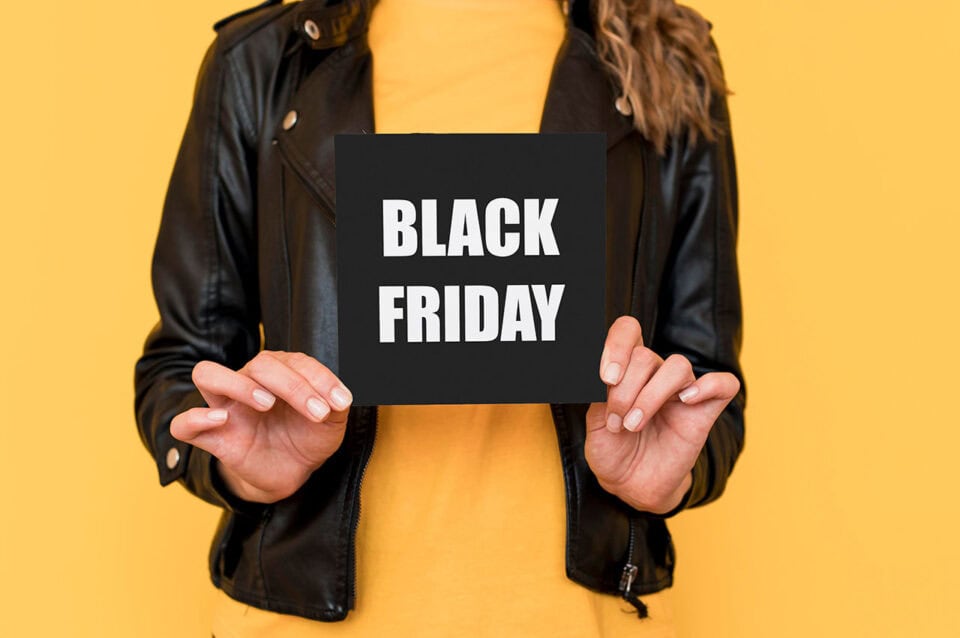 Eine Person in schwarzer Lederjacke und gelbem Hemd hält ein schwarzes Schild mit der Aufschrift "BLACK FRIDAY" in weißen Großbuchstaben auf gelbem Hintergrund - perfekt für Dienstleister, die neue Kunden anlocken wollen.
