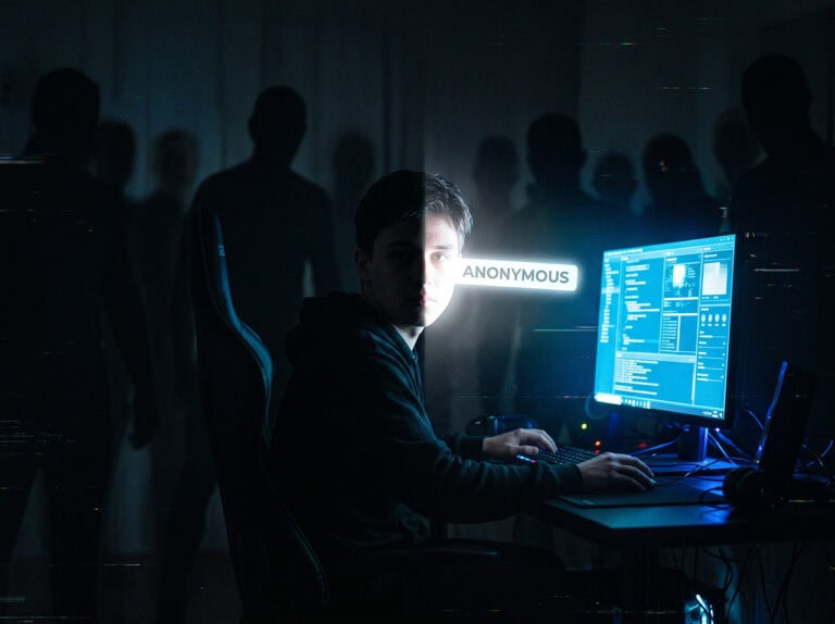 Eine Person sitzt in einem dunklen Raum an einem Computer mit mehreren Monitoren, umgeben von schattenhaften Gestalten, unter dem beleuchteten Wort "ANONYMOUS" - ein starker Kontrast zur Idee der Klarnamenpflicht und der Regel gegen die Verwendung falscher Identitäten.